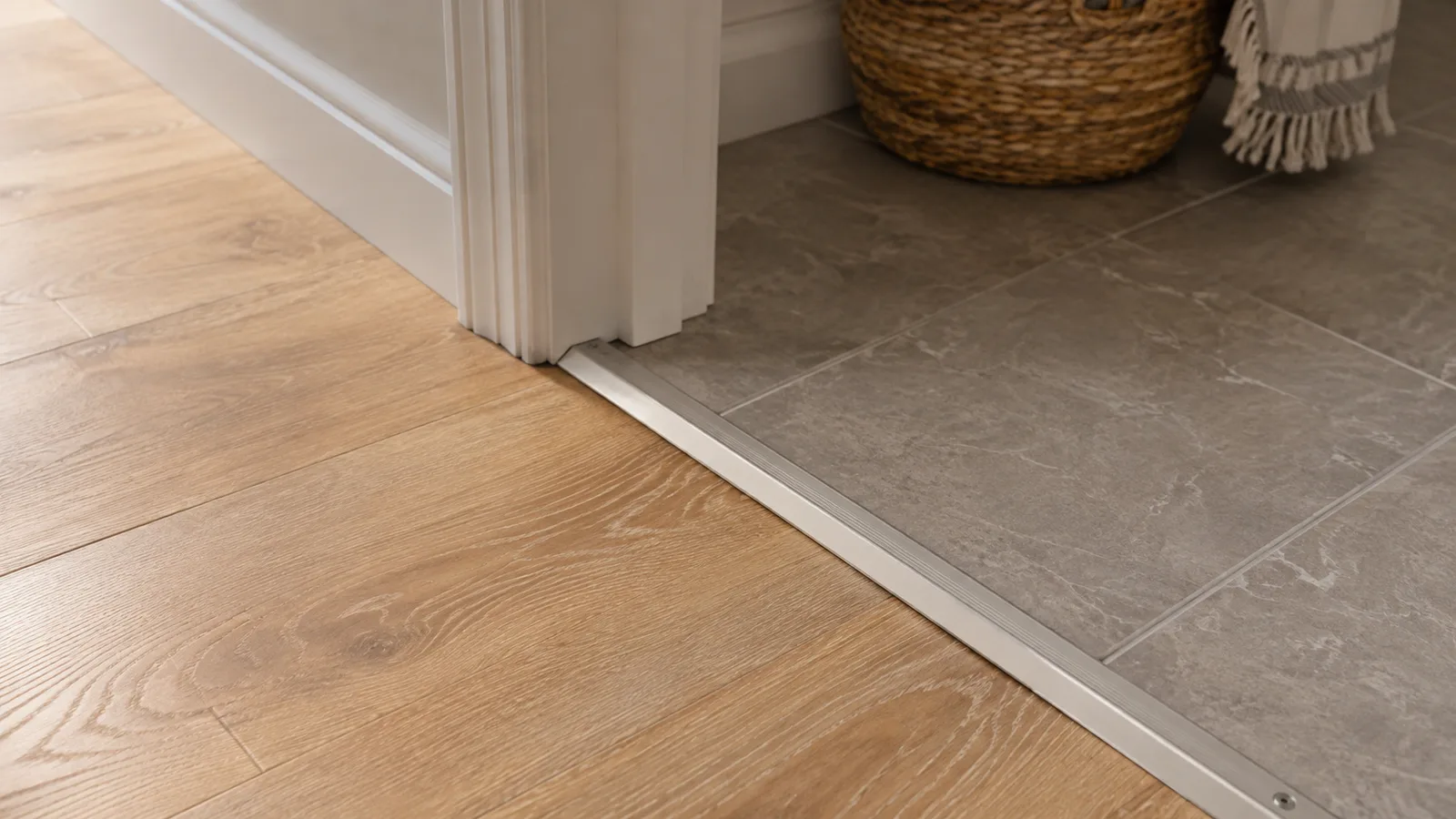 Light oak plank flooring meeting gray tile at a doorway with a metal T-molding