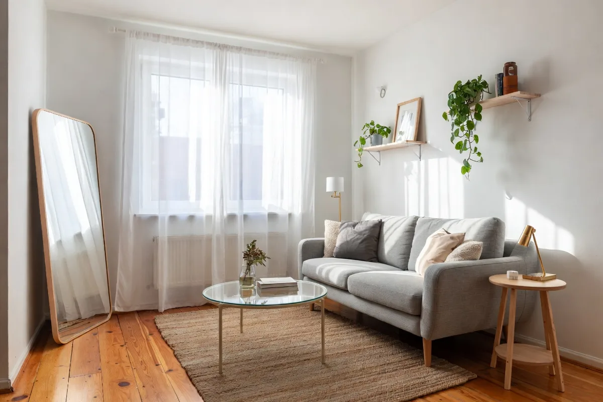 Small apartment living room decorated with a compact gray sofa, glass coffee table, sheer curtains, and large mirror for a spacious feel
